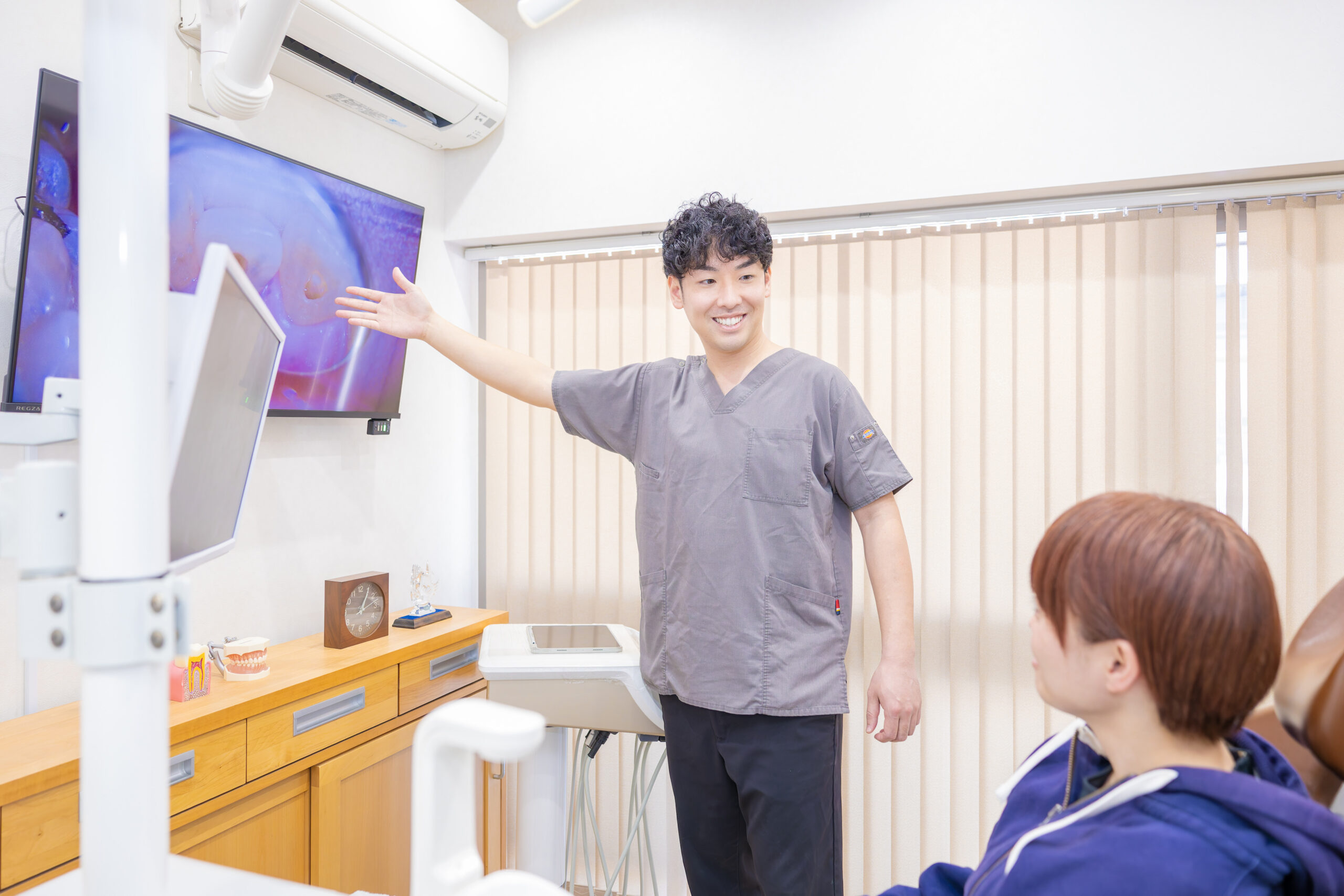 Dentist in gray scrubs explains a dental image on a large screen to two patients in a bright clinic.
