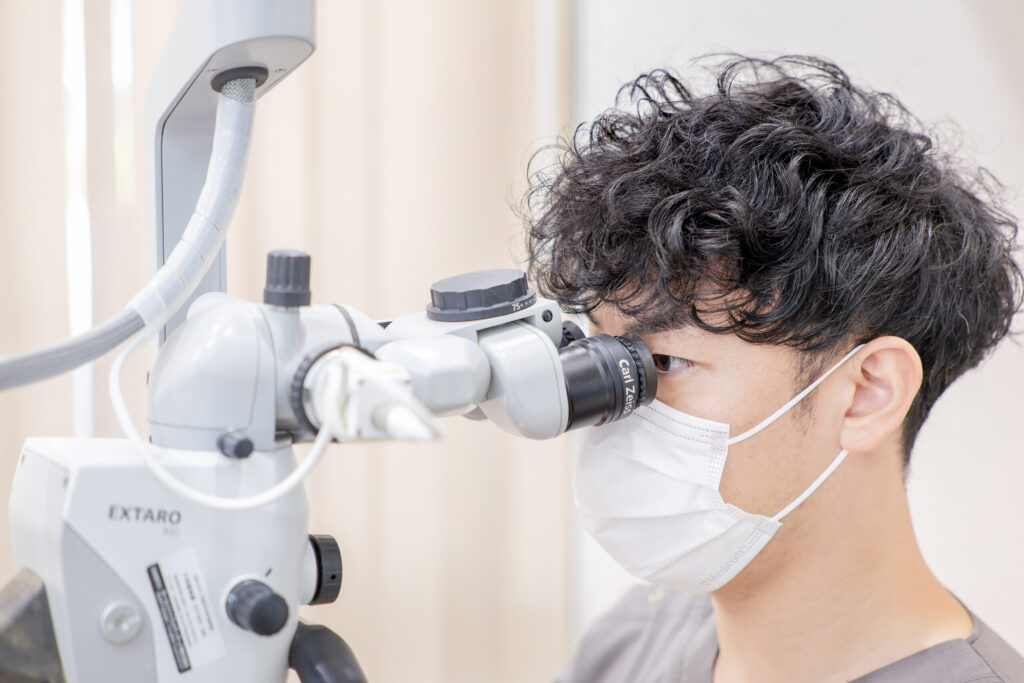 Person wearing a medical mask looks into a stereo microscope for a clinical examination in a lab setting, facing right side of image.