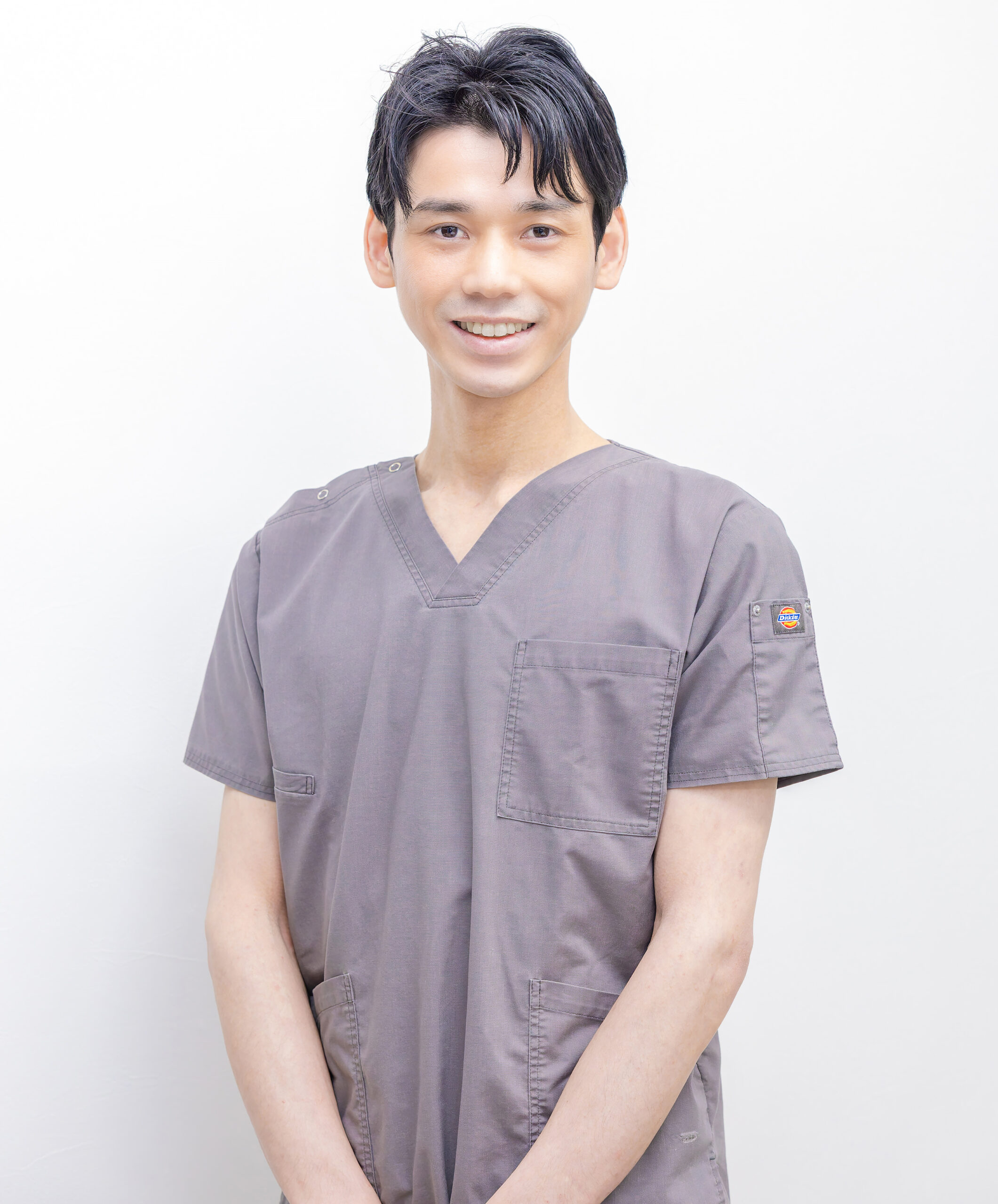 Young man in gray medical scrubs smiling at the camera against a plain white background.