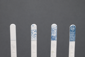 Four white, vertical test strips with blue staining on the tips, arranged left to right against a dark background (increasing stain intensity).
