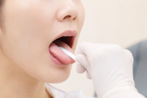 Close-up of a person’s mouth as a gloved clinician inserts a white oral swab onto the tongue for a medical test.