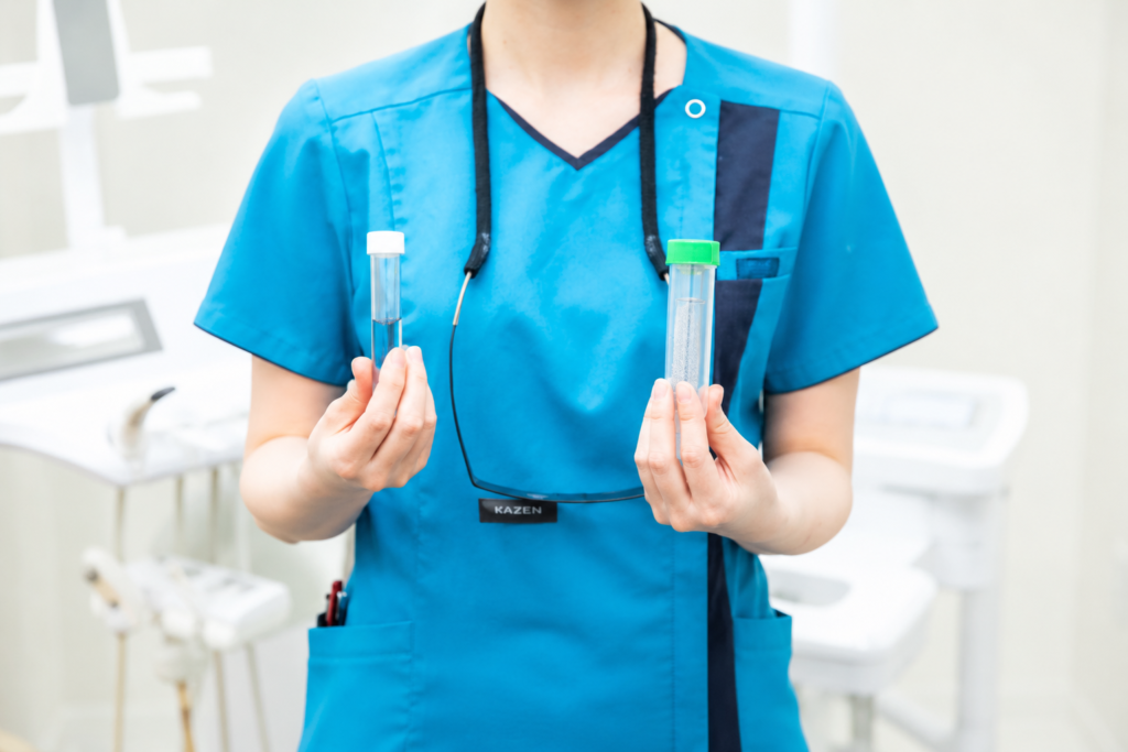 Medical professional in blue scrubs holds two specimen tubes with caps (white and green) in a clinical setting.