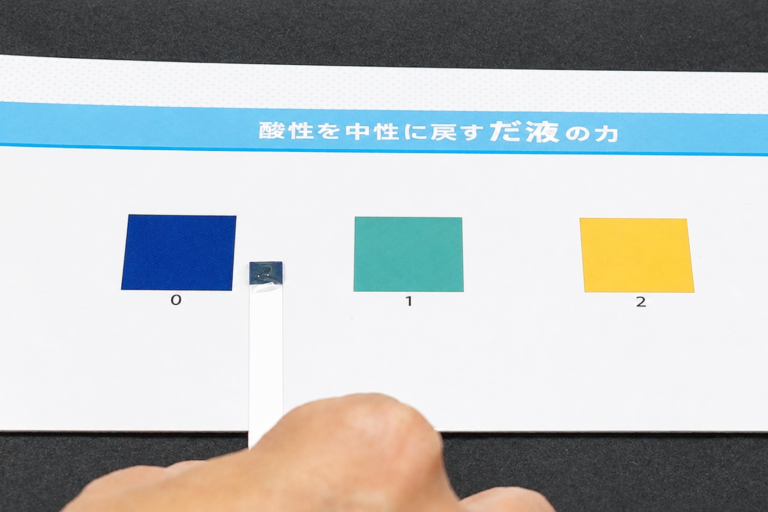 Color reference card showing three swatches labeled 0 (blue), 1 (teal), and 2 (yellow). A hand holds a small shiny square near swatch 0 as it’s being placed or adjusted.